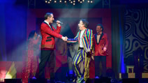 CIRQUE - The Greatest Show cast on stage with a man in a rainbow suit looking surprised as he shakes the hand of a man in a red sequin jacket who is singing to him