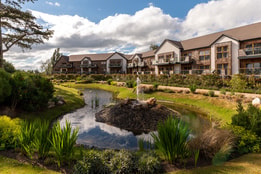 Serene landscape at Alvaston Hall featuring modern accommodations beside a tranquil pond with a fountain, surrounded by lush greenery and blue skies.