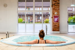 A woman relaxes the bubble pool at Alvaston Hall Hotel, facing large windows that overlook a green garden area. A clock is visible on the wall