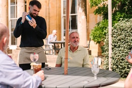 A waiter in a black shirt shakes a cocktail shaker at an outdoor table, while guests enjoy drinks under a sunny sky a Cricket St. Thomas