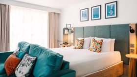 Contemporary hotel bedroom featuring a king-size bed with a padded headboard, decorative patterned cushions and a teal sofa at the foot of the bed. Large windows with floor-length curtains fill the room with natural light.