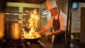 Chef in a professional kitchen cooking with a pan over open flames on a gas stove, with a large pot beside it and industrial kitchen equipment in the background.