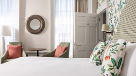 A bright bedroom seating area with two upholstered chairs and a small round table beside a window, next to a neatly made bed with botanical cushions and patterned wallpaper.
