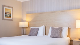 Modern hotel bedroom with a large white bed, wooden headboard, textured cushions, soft bedside lighting, and neutral-toned walls with framed artwork.