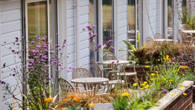 Outdoor garden seating area with round tables and chairs set among colourful flowers and plants, alongside a row of light blue lodge-style buildings with glass doors.
