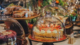 Display of cakes and pastries arranged on wooden stands and covered with glass domes, including a sponge cake decorated with strawberries and cream, pastries drizzled with icing, and other desserts set against a softly lit café background with greenery and blurred lights.