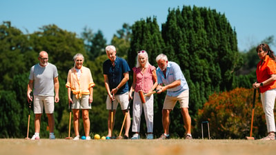 A group of six individuals wearing casual summer clothing stand on a grassy field, preparing to play croquet against a sunny backdrop.