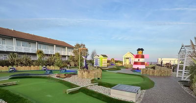 A colorful mini-golf course featuring a striped lighthouse, beach huts, and lush greenery, with playful blue and green pathways.