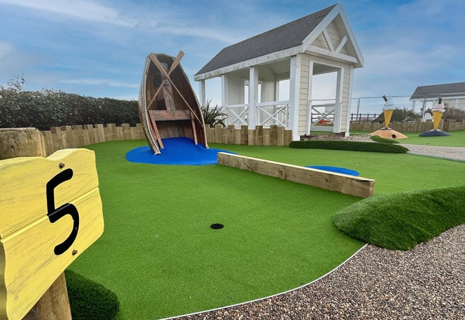 A colorful mini-golf course featuring a boat obstacle, a small blue pond, and a white gazebo, with a visible hole number sign.
