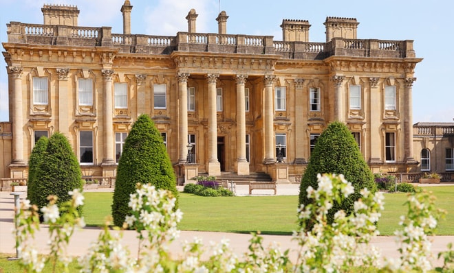 The main Heythrop Park hotel building surrounded by manicured hedges and blooming flowers on a sunny day.