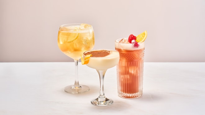 Three colorful cocktails on a marble surface: a bubbly drink with lemon, a creamy one with chocolate, and a fruity mix with raspberries.