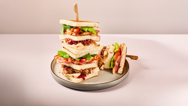 A tall stack of layered sandwiches with lettuce, tomatoes, and meat, served on a round plate against a soft pink background.