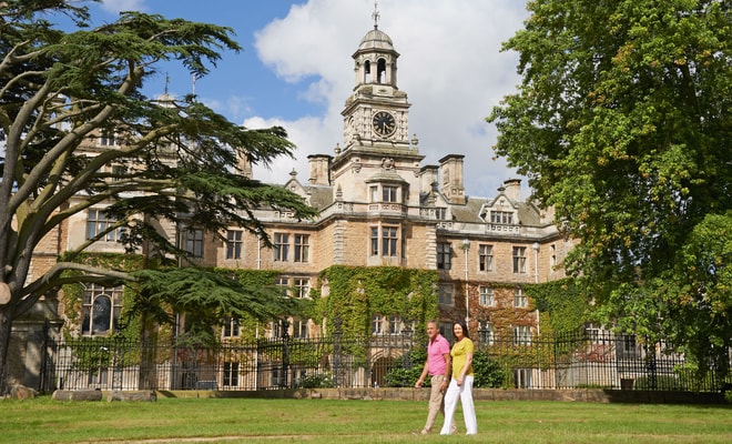 A couple strolls across a lush green lawn in front of a grand historic mansion, adorned with ivy and surrounded by large trees at Thoresby Hall