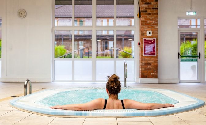 A woman relaxes the bubble pool at Alvaston Hall Hotel, facing large windows that overlook a green garden area. A clock is visible on the wall