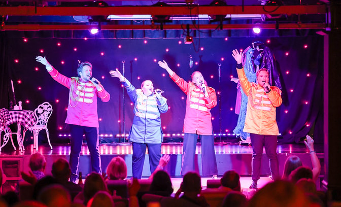 Four members of the Warner Theatre Company at Bembridge Coast performing on stage, with guests waving and joining in.