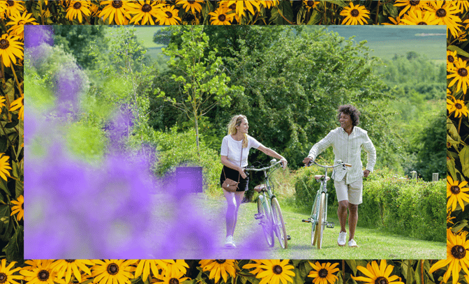 A woman and a man walk with bicycles through a vibrant garden filled with lush greenery and colorful flowers.