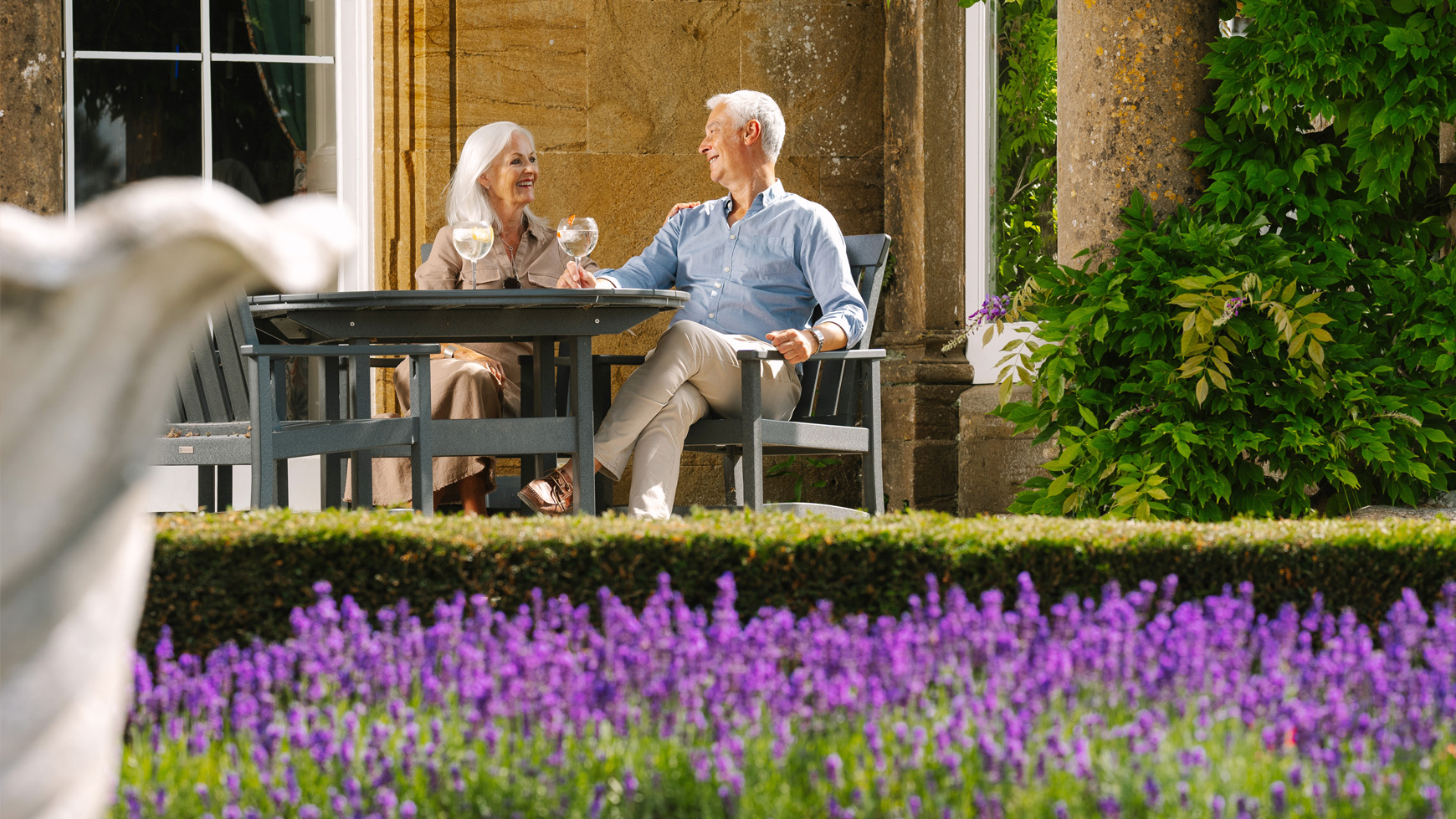 A couple seated at a garden table at Cricket St. Thomas with glasses of wine, surrounded by vibrant purple flowers and lush greenery.