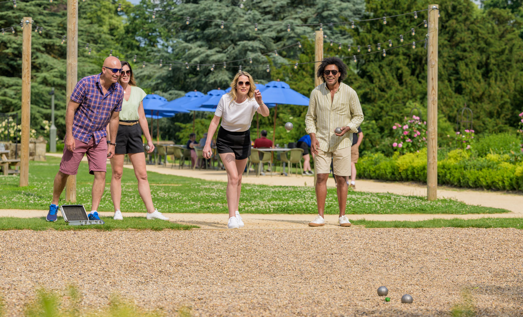 A group of four people plays p&eacute;tanque on a sunny day at Studley Castle, with green trees and blue umbrellas in the background.