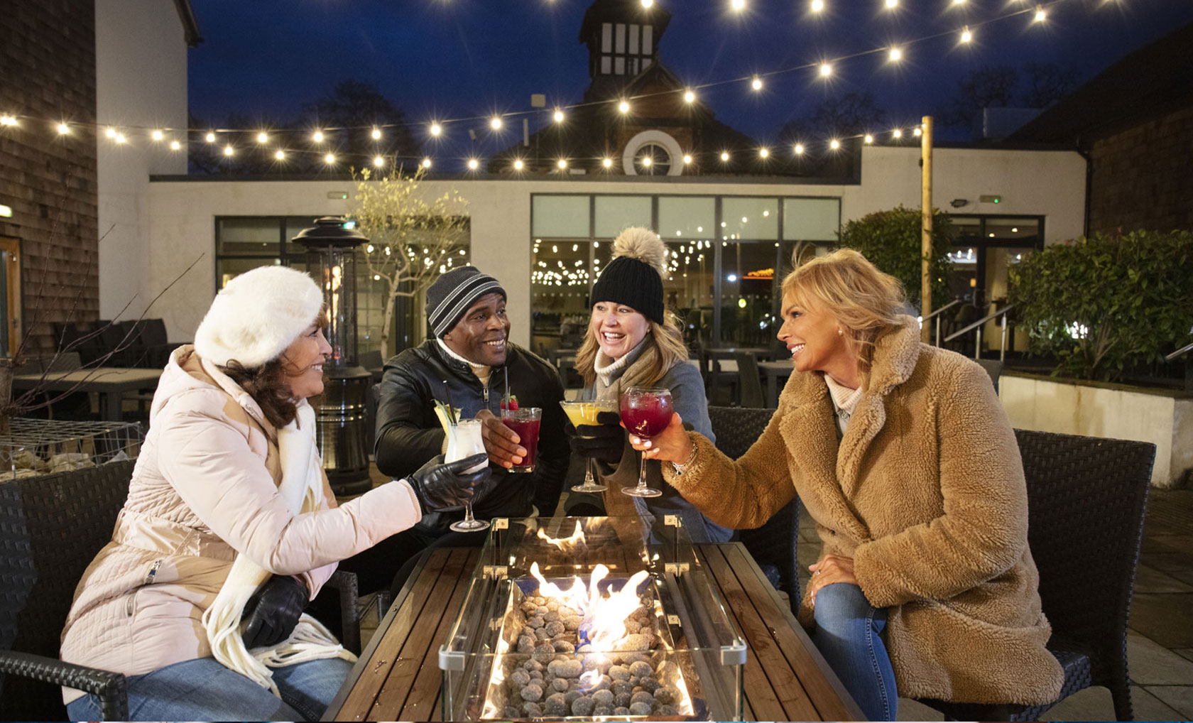 A group of friends enjoys colorful cocktails around a fire pit, under string lights, on a cozy evening outdoors.