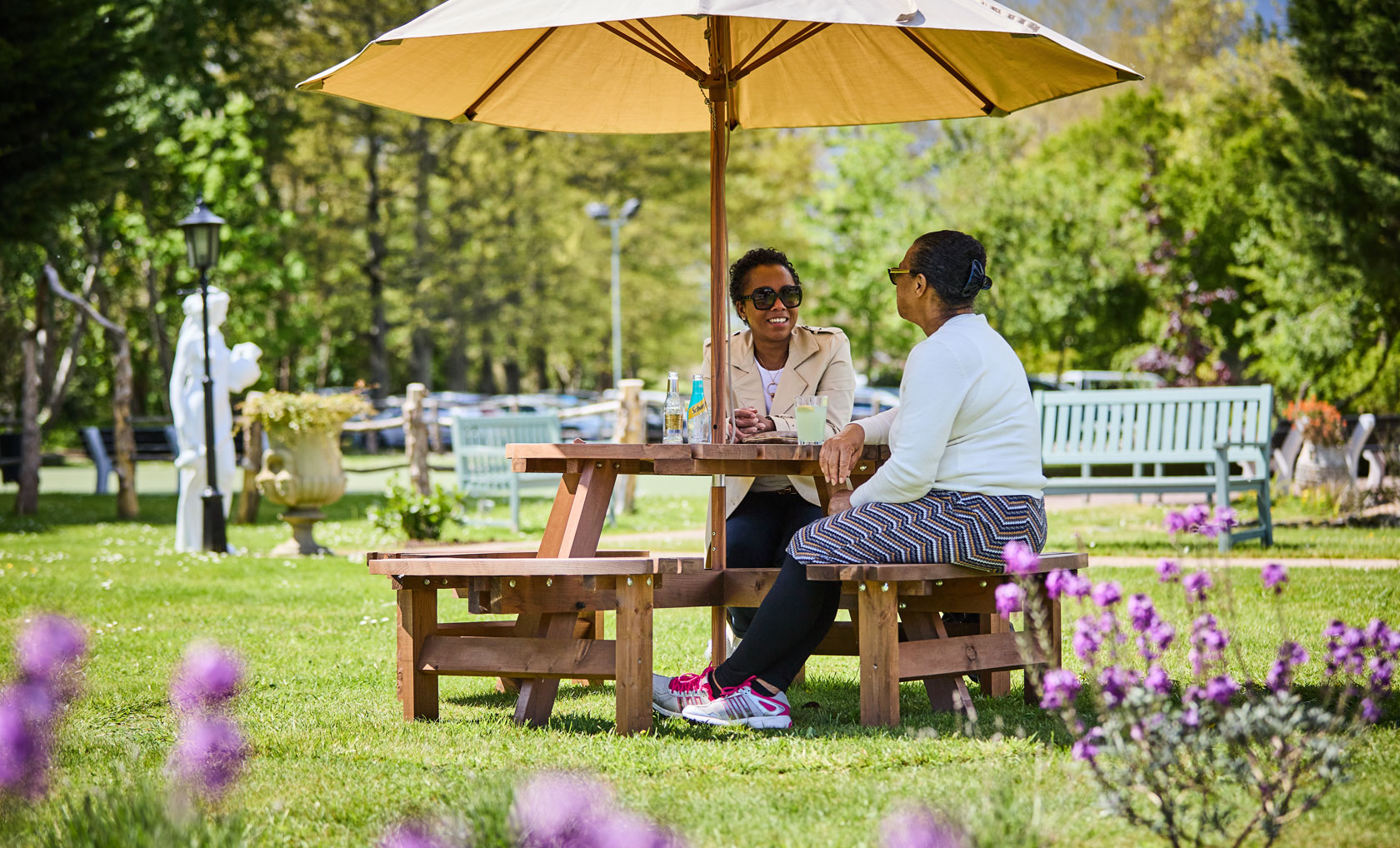 Two women sit at a wooden picnic table under a large umbrella, surrounded by lush greenery and vibrant flowers in a sunny park.